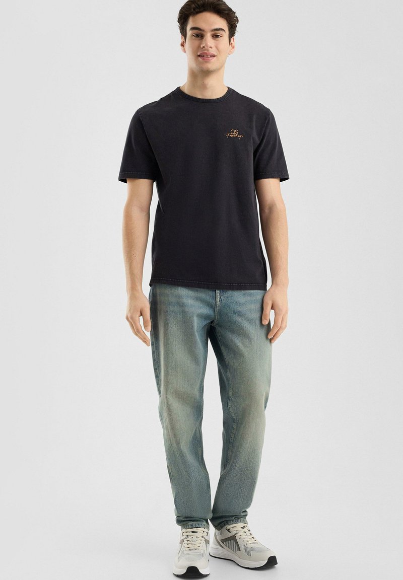 Zwarte katoenen t-shirt met korte mouwen en een ronde hals, voorzien van een klein gouden geborduurd logo. Gepaarde met lichtblauwe denim jeans.