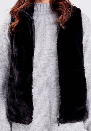 Kvinde iført en lysegrå ribstrikket sweater under en åben sort fluffy vest, der viser midt på kroppen og en del af bølget brunt hår.