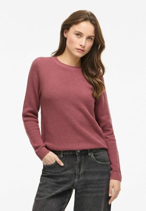 Femme aux longs cheveux bruns portant un pull texturé mauve et un jean noir, posant avec une main dans la poche sur un fond blanc.