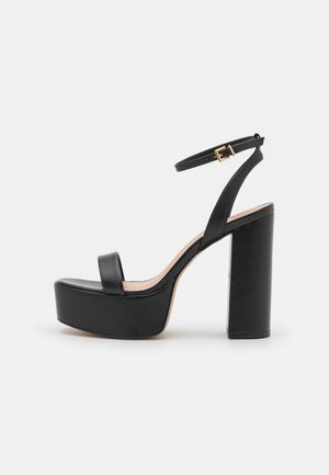 Μαύρες ψηλοτάκουνες σανδάλια με παχιά πλατφόρμα, λεία υφή δέρματος, λουράκι στον αστράγαλο με χρυσό κούμπωμα και τετράγωνη block τακούνι.