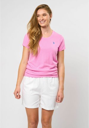 Tricou din bumbac roz deschis, cu guler rotund și mâneci scurte, cu un mic logo albastru pe piept, purtat cu pantaloni scurți albaștri cu talie elastică.