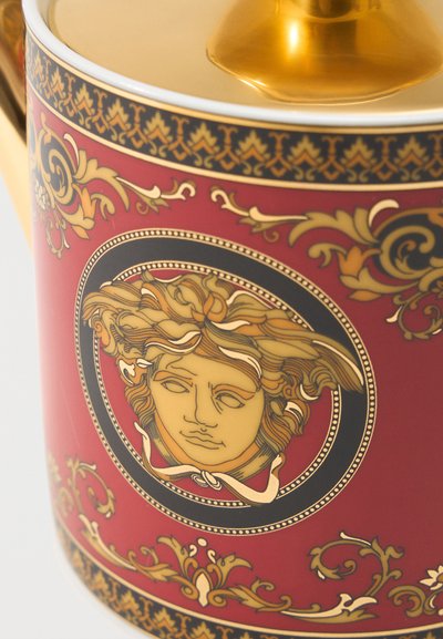 Gros plan sur une céramique rouge et dorée avec des motifs baroques ornés et un emblème central de tête de Méduse en tons noirs et dorés.