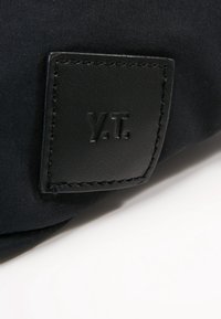 Musta kangaskassi, jossa on tekstuurinen nahkapatch, jossa on kohokuvioidut aloitteet "Y.T." ja kontrastiväriset ompeleet reunoilla.