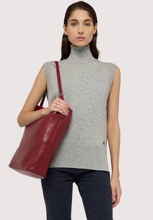 Grå ærmeløs rullekravetrøje med ribstrikket struktur og en rød læder tote taske, båret af en model. Mørke bukser kompletterer outfittet.