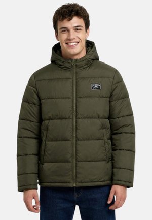 Joven sonriente que lleva una chaqueta acolchada con capucha de color verde oliva con cremallera frontal y bolsillos laterales sobre un fondo liso.