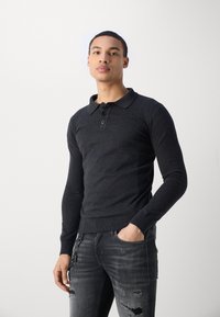 Mørkegrå langærmet polo skjorte med krave, tre knapper, tætsiddende design og ribbede manchetter, parret med slidte sorte jeans.