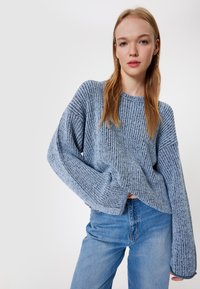 Jasnoniebieski sweter dzianinowy z szerokimi rękawami i fakturowaną powierzchnią. W połączeniu z niebieskimi dżinsami podkreślającymi luźny krój.