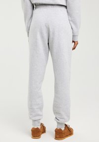 Osoba ubrana w jasnoszare spodnie dresowe i pasującą bluzę, pokazana od tyłu, w brązowych butach codziennych na jednolitym tle.