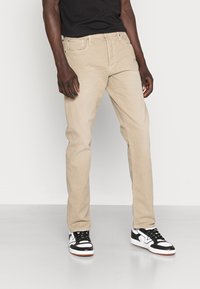 Chinos bege com corte reto, apresentando bolsos frontais e um passante de cinto visível. Combinados com ténis em preto e branco.