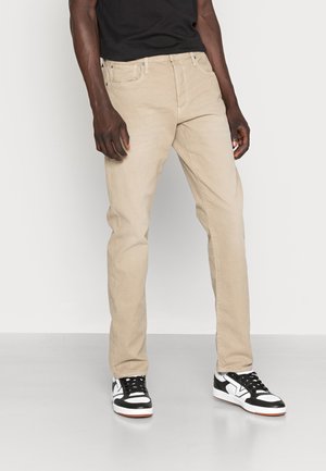 Chinos bege com corte reto, apresentando bolsos frontais e um passante de cinto visível. Combinados com ténis em preto e branco.