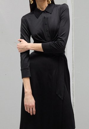 Kobieta ubrana w czarną, długorękawową sukienkę z guziczkami, z kołnierzykiem i paskiem w talii, z dużymi złotymi kolczykami, z rękami skrzyżowanymi na piersi.