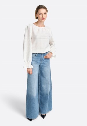 Weiße strukturierte Bluse mit Puffärmeln, kombiniert mit hellblauen weitgeschnittenen Jeans. Schwarze spitze Absätze runden das Outfit ab.
