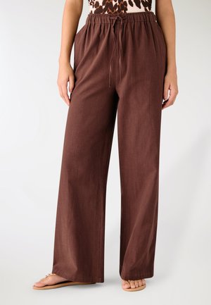 Pantaloni marroni a gamba larga con vita elasticizzata e coulisse indossati da una persona in sandali, su uno sfondo neutro.