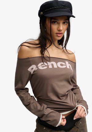 Ung kvinde iført en brun off-shoulder top med "Bench" logo og en sort kasket, med langt mørkt hår, der falder over skuldrene.
