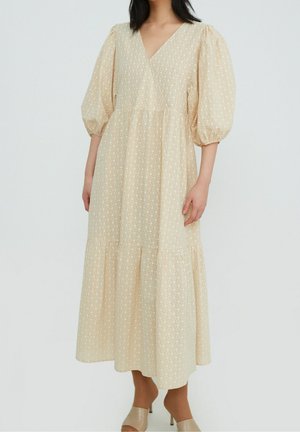 Kvinde iført en beige midikjole med puffede ærmer, V-udskæring og et diskret blomsterprint, kombineret med beige åbne hæle.