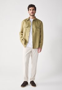 Jachetă cămilie de un verde măsliniu, cu două buzunare la piept, închidere cu buton și o croială lejeră, asortată cu pantaloni crem și pantofi negri.
