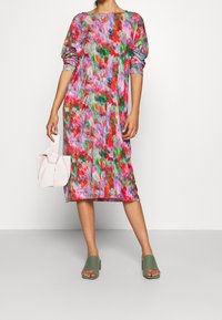 Bunte Blumen-Midi-Kleid mit puffäromel-Ärmeln in den Farbtönen Pink, Grün und Rot. Begleitet von einer kleinen hellrosa Tasche und grünen Blockabsatzsandalen.