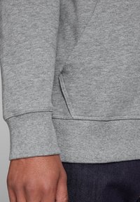 Sudadera gris de tejido suave y texturizado. Presenta un bolsillo canguro y puños acanalados. Mano visible, descansando sobre una superficie oscura.