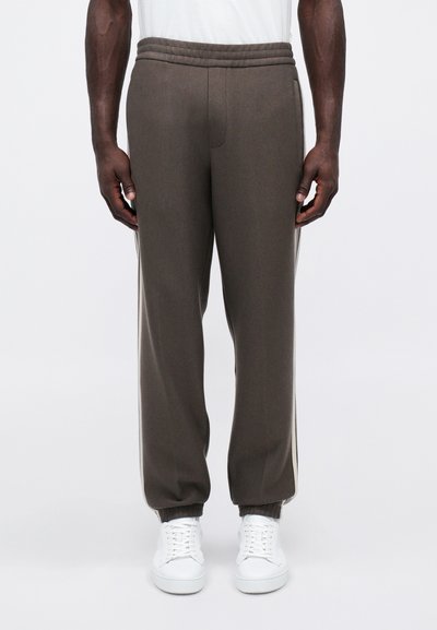 Άνδρας που φοράει καφέ στενά παντελόνια με ελαστικές μανσέτες και λευκά αθλητικά παπούτσια, στέκεται μπροστά από λευκό φόντο.