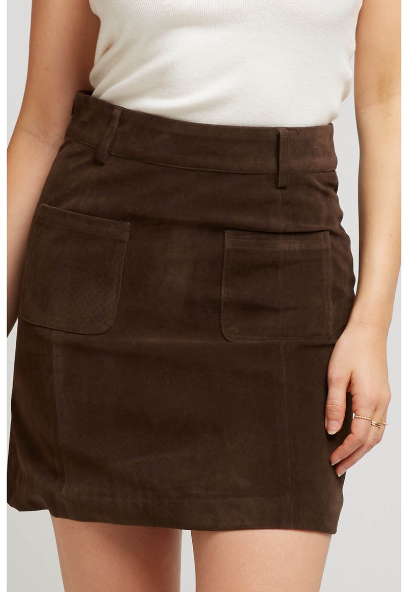 Femme portant une mini-jupe en suedine marron foncé avec des poches avant et un haut sans manches blanc, la main reposant près de la jupe.