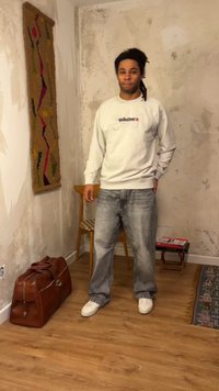 Homme avec des dreadlocks se tient dans une pièce usée, portant un sweat blanc Quiksilver, un jean délavé et des baskets blanches près d'une chaise et d’un sac en cuir.