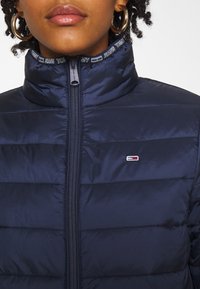 Gros plan sur une personne portant une veste matelassée bleu marine avec zip et le logo Tommy Jeans près de la poitrine, de grandes boucles d'oreilles visibles.