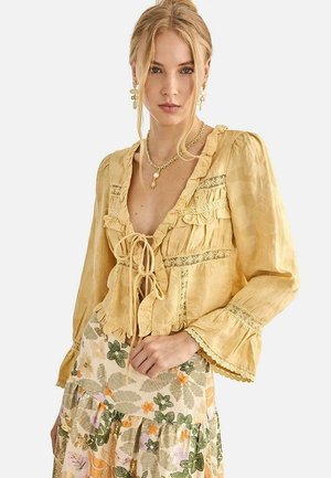 Mujer rubia vistiendo una blusa amarilla con volantes y detalles de encaje, junto con una falda de estampado floral, complementada con joyería dorada.