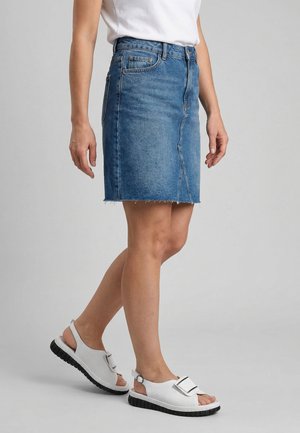 Frau trägt einen blauen Jeansrock, ein weißes T-Shirt und weiße offene Sandalen mit schwarzen Sohlen, steht vor einem schlichten grauen Hintergrund.