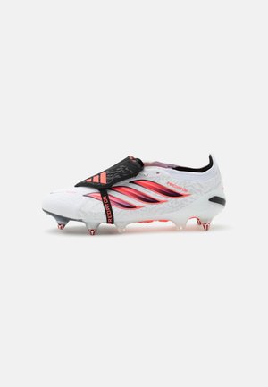 Biały korki piłkarskie Adidas Predator z czerwonymi paskami, czarnym klapanym językiem i metalowymi kołkami na podeszwie.