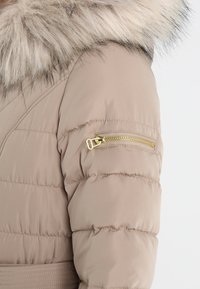 Prošívaná bunda světle béžové barvy s kapucí lemovanou kožešinou. Má zipovou kapsu na rukávu se zlatým kováním a prošívané detaily.
