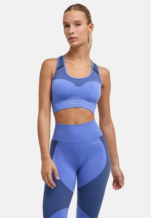 Vrouw die staat, gekleed in een bijpassende blauwe naadloze sportbeha en hooggetaillede legging met donkerblauwe accenten op een witte achtergrond.
