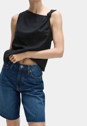 Vrouw met zwart mouwloos topje met geknoopte schouderdetail en blauwe denim knielange korte broek, die haar shirt met één hand rechtzet.