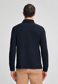 Polo a maniche lunghe da uomo in nero, con una texture morbida, colletto e polsini a coste, indossato con pantaloni beige, mostrando la vista posteriore.