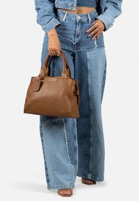 Brun læderhåndtaske med to håndtag, rektangulær form, tekstureret finish og en lille fastgjort mærkat. Bæres med lyseblå vidde jeans.