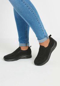 Chaussures noires en tricot à enfiler avec une tige texturée, une semelle flexible et une languette au talon, portées avec un jean bleu retroussé.
