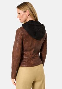 Braune Lederjacke mit einer schwarzen Stoffkapuze, taillierter Schnitt und sichtbaren Nähdetails, kombiniert mit beigen Hosen.