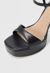 Sandalo platform nero con punta quadrata, tomaia sintetica liscia e due ampie fascette. Presenta una soletta beige e dettagli metallici sulla suola.