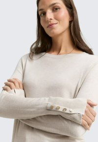 Beige langesweater met lange mouwen, gemaakt van zacht materiaal, met ribgebreide manchetten en vier decoratieve gouden knopen, evenals uitsparingen op de mouwen.
