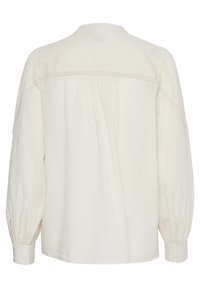 Blusa color crema chiaro con maniche lunghe a sbuffo, tessuto testurizzato con piccoli pois, dettagli in pizzo lungo lo scollo e polsini con bottoni.