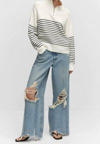 Kremowo-czarny sweter w paski z wysokim kołnierzem, w połączeniu z jasnoniebieskimi jeansami o szerokich nogawkach, z dużymi przetarciami i postrzępionymi brzegami.