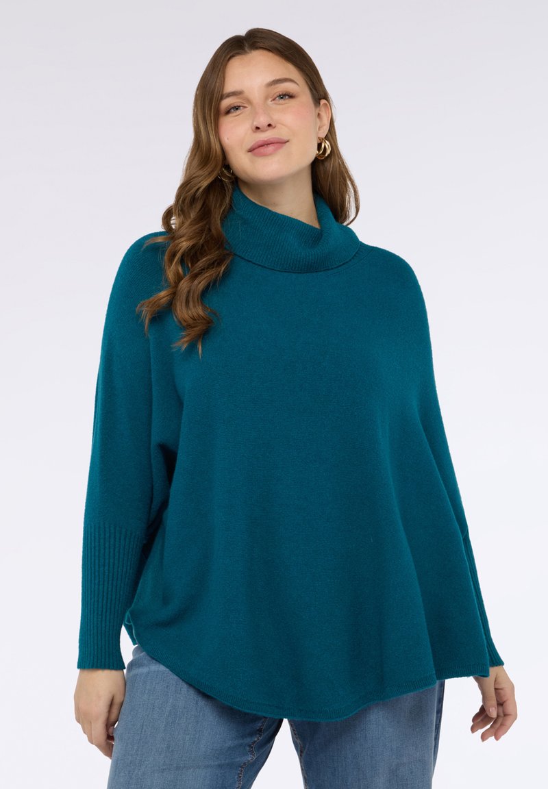 Maglione oversize color verde petrolio con collo alto e polsini a coste, realizzato in materiale morbido. Vestibilità ampia e orlo arrotondato, indossato sopra jeans blu. Sfondo semplice.