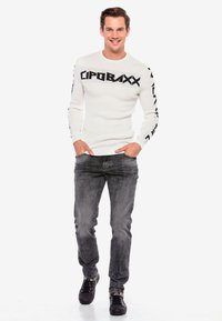 Weißer, gerippter Pullover mit schwarzem Grafiktext auf den Ärmeln, kombiniert mit grauen gemusterten Jeans und schwarzen Sneakern. Modell steht mit den Händen in den Taschen.