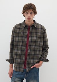 Camicia verde a quadri con colletto, chiusura a bottoni e maniche arrotolate, indossata sopra una camicia bordeaux, abbinata a jeans azzurri chiari.