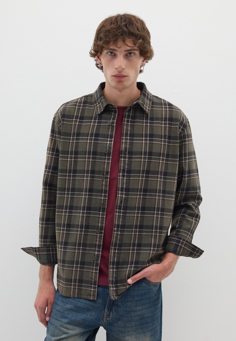 Camicia verde a quadri con colletto, chiusura a bottoni e maniche arrotolate, indossata sopra una camicia bordeaux, abbinata a jeans azzurri chiari.