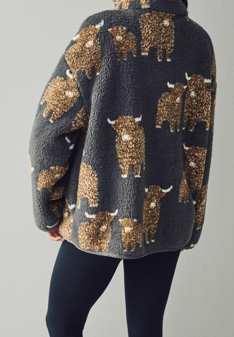 Veste polaire gris foncé avec un motif de vache des Highlands marron et beige, portée par une personne en pantalon noir, vue de dos.
