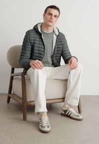 Grå pufferjakke, grå sweater, hvide bukser og multikolorede sneakers. Model sidder på en træstol mod en ensfarvet baggrund.