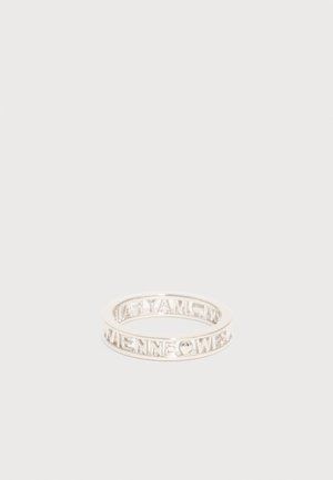 Bague fine en argent avec des lettres découpées formant une phrase enroulée autour de l'anneau, posée sur une surface blanche unie.