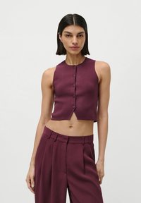 Žena s krátkymi tmavými vlasmi v bezrukávovom bordovom crop topi s gombíkmi a zodpovedajúcich vysokých nohaviciach, stojaca pred jednoducho bielym pozadím.