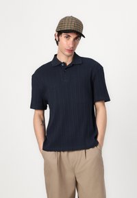 Navy ribbed polo shirt with a collar and buttons, paired with beige trousers. The model wears a plaid cap with a rounded peak.