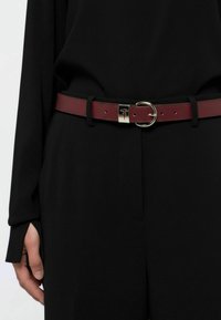 Cinturón de cuero burdeos con hebilla plateada, con un diseño circular y agujeros ajustables, usado sobre pantalones de tela negra.